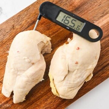 Two pieces of poached chicken on a wooden cutting board with a thermometer sticking out of one of them registering at 165.