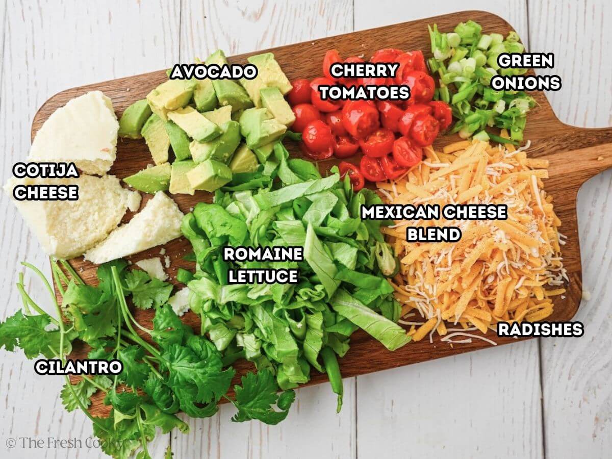 Labeled ingredients for toppings tostadas, L-R Cheese, avocado, cherry tomatoes, green onion, Mexican cheese, romaine lettuce, cilantro.