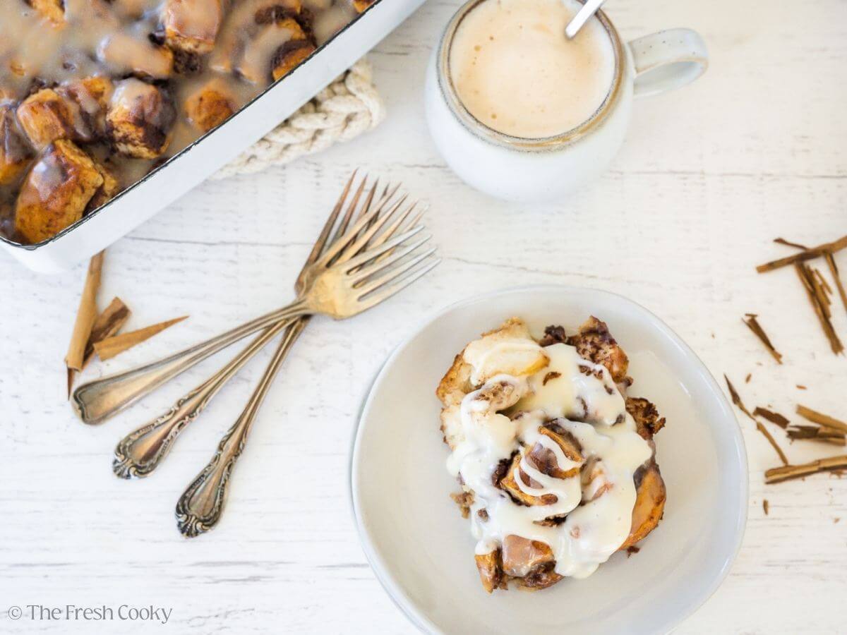 A serving of cinnamon roll casserole with frosting near the pan, and a cup of coffee.