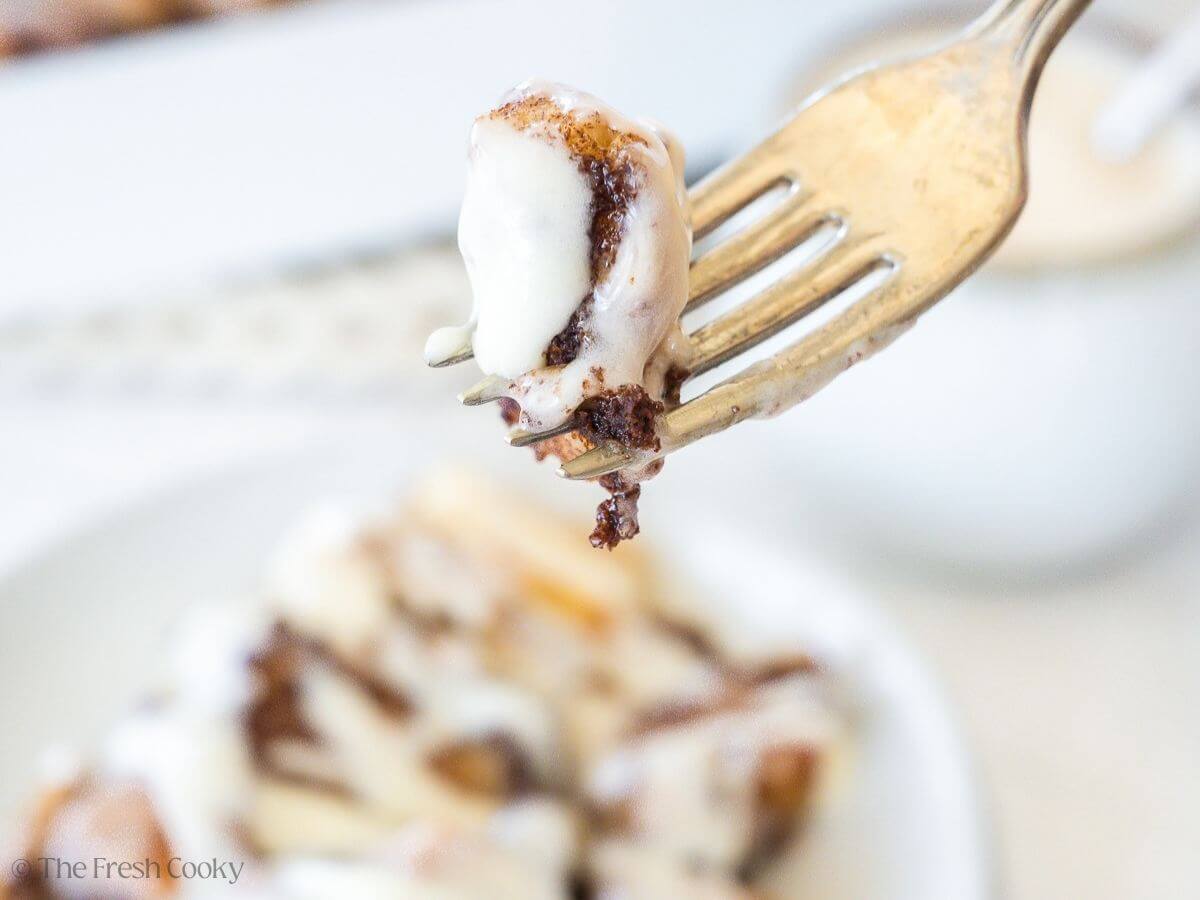 A bite on a fork of gooey cinnamon roll casserole, with the serving in background on a white plate.