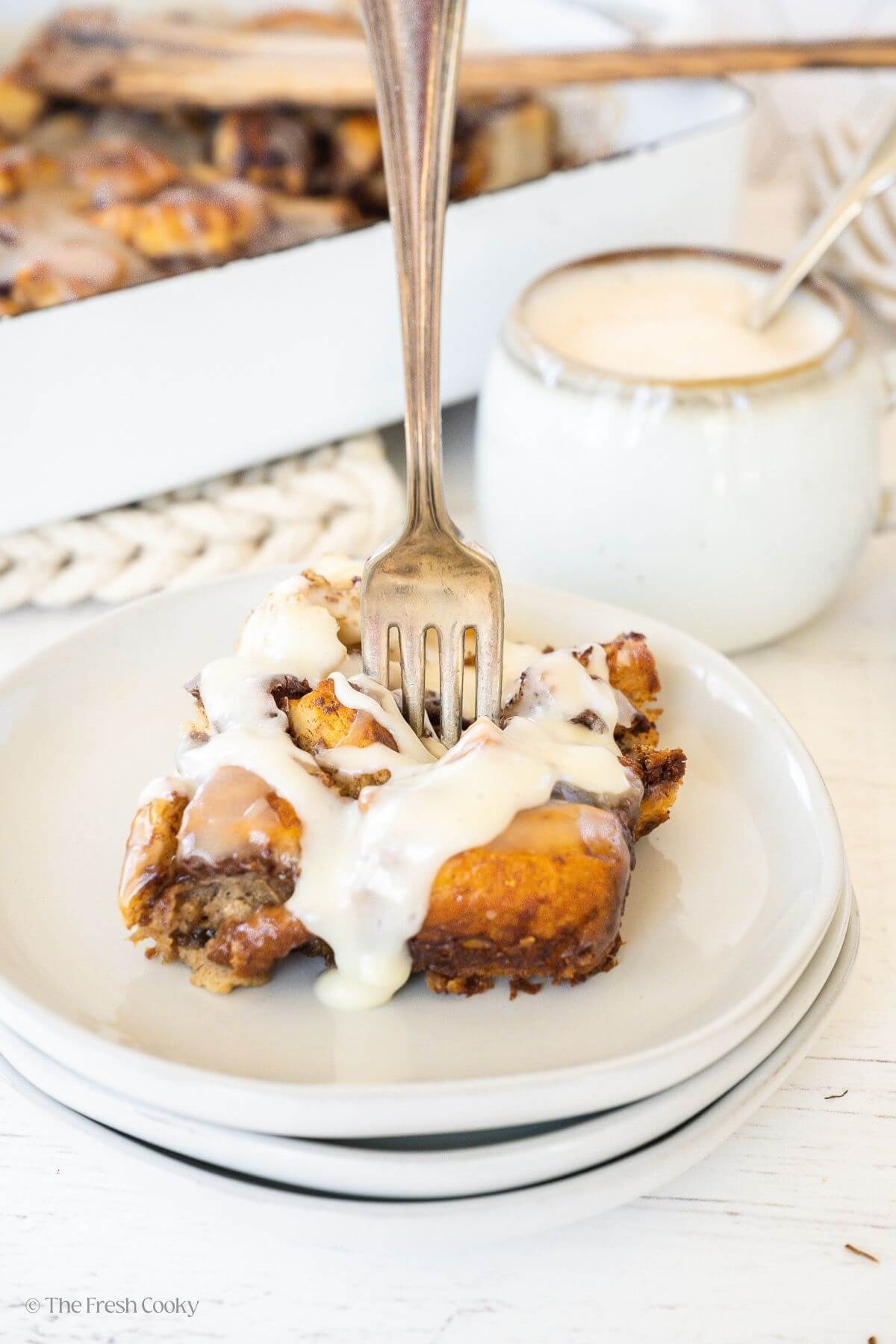 A serving of cinnamon roll casserole topped with frosting on a white plate, with a cup of coffee behind.