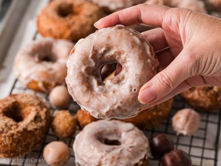 Hand holding a glazed donut over more donuts on a wire rack.