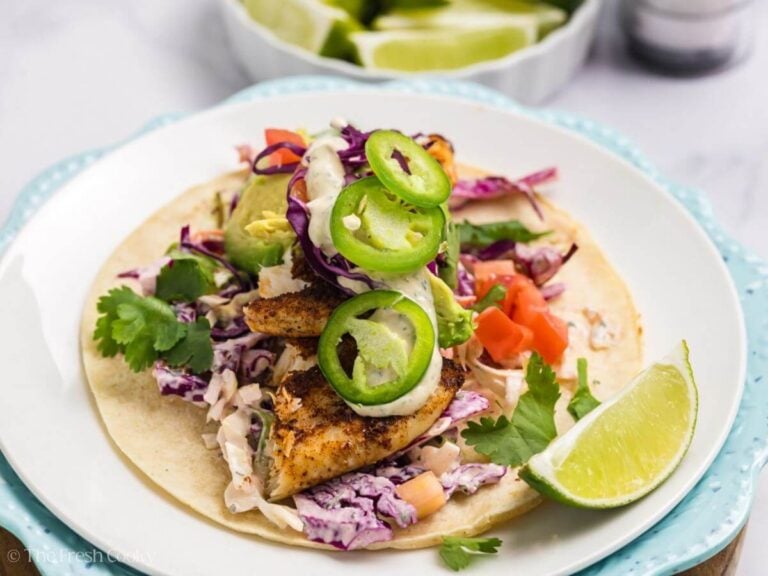 Side view of a fish taco with slaw, avocado, jalapenos, and a lime wedge on the plate.