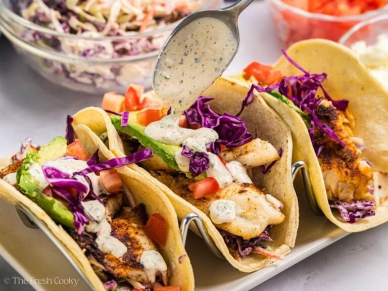 Three tilapia fish tacos in a taco holder, drizzling sauce on top with a spoon.