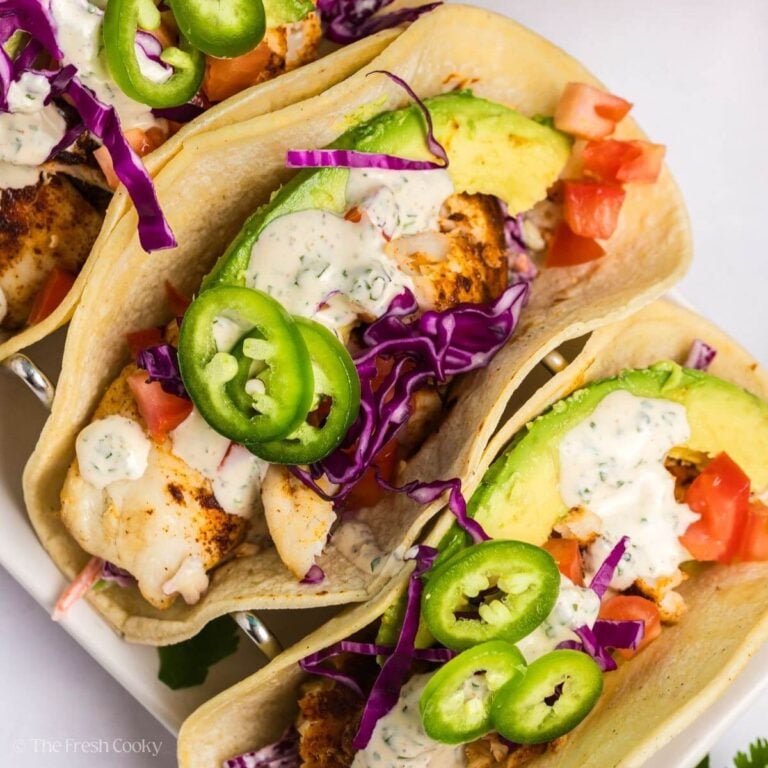 Three tilapia fish tacos all topped with sauce, avocado, tomatoes and jalapenos.
