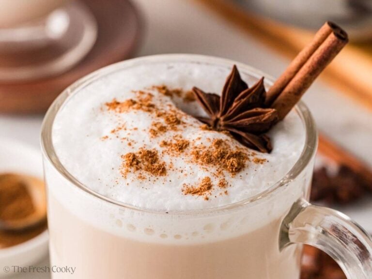 Close up of the top of a chai tea latter with cinnamon sprinkled on top, next to a whole anise and cinnamon stick.