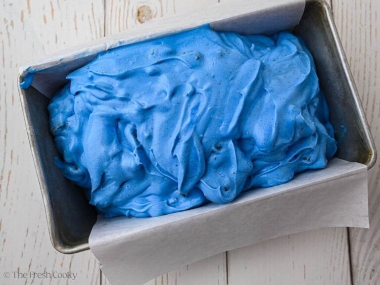 Blue ice cream mixture with cookies in it poured into a loaf cake lined with parchment paper.