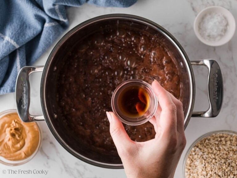 Sugar and cocoa mixture boiling in a saucepan with a hand holding a small bowl of vanilla.