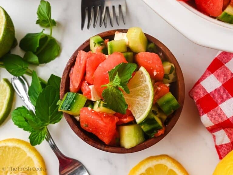 Watermelon feta cucumber salad in a wooden bowl with fresh lemon, mint and lime nearby.