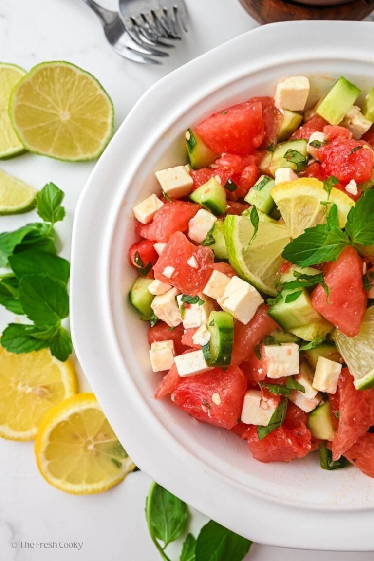 Overhead shot of a large bowl of watermelon cucumber salad next to slices of lemons and limes.