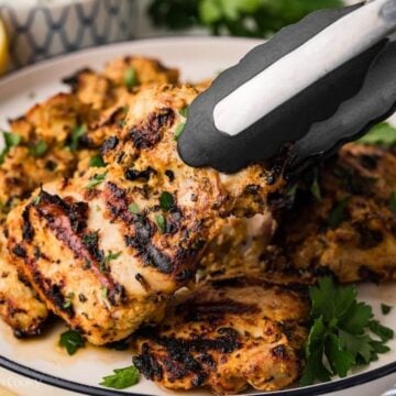 Tongs grabbing a piece of grilled Greek chicken off a platter.