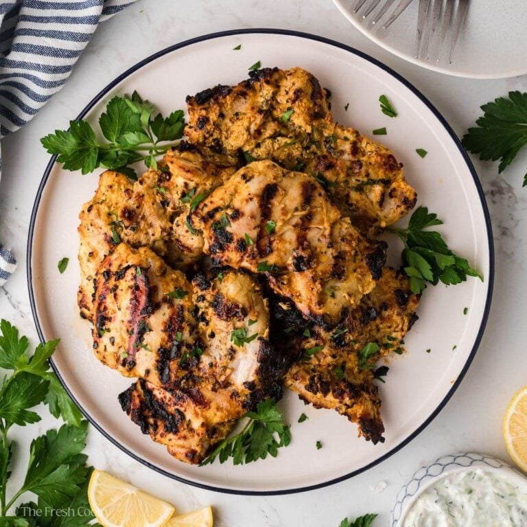 Looking down on a plate of grilled chicken thighs that have been marinated in a Greek yogurt marinade, surrounded by parsley and lemons.
