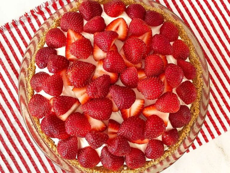Looking down on a beautiful strawberry no-bake cheesecake, topped with fresh halved strawberries in a graham cracker crust, on a striped red and white placemat.
