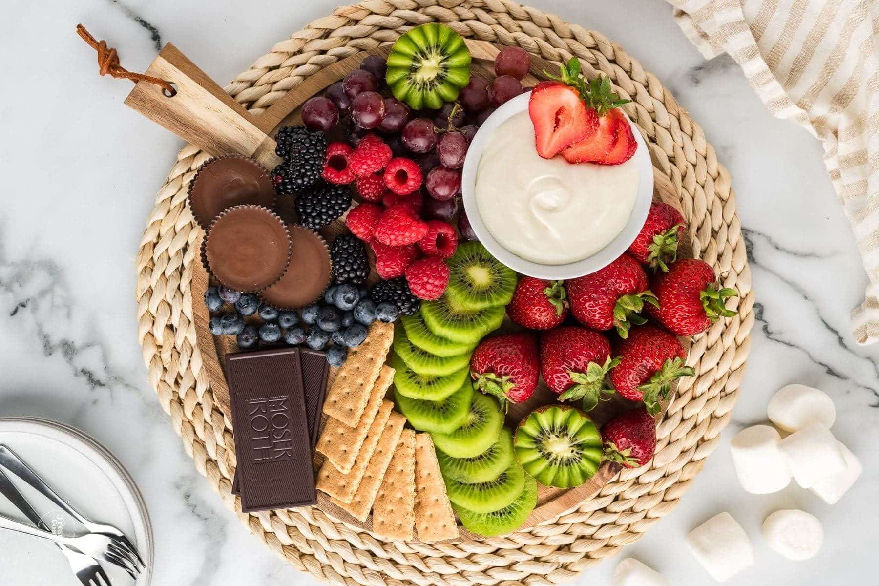 Platter with fresh fruits, crackers and chocolate surround a bowl of marshmallow fruit dip.