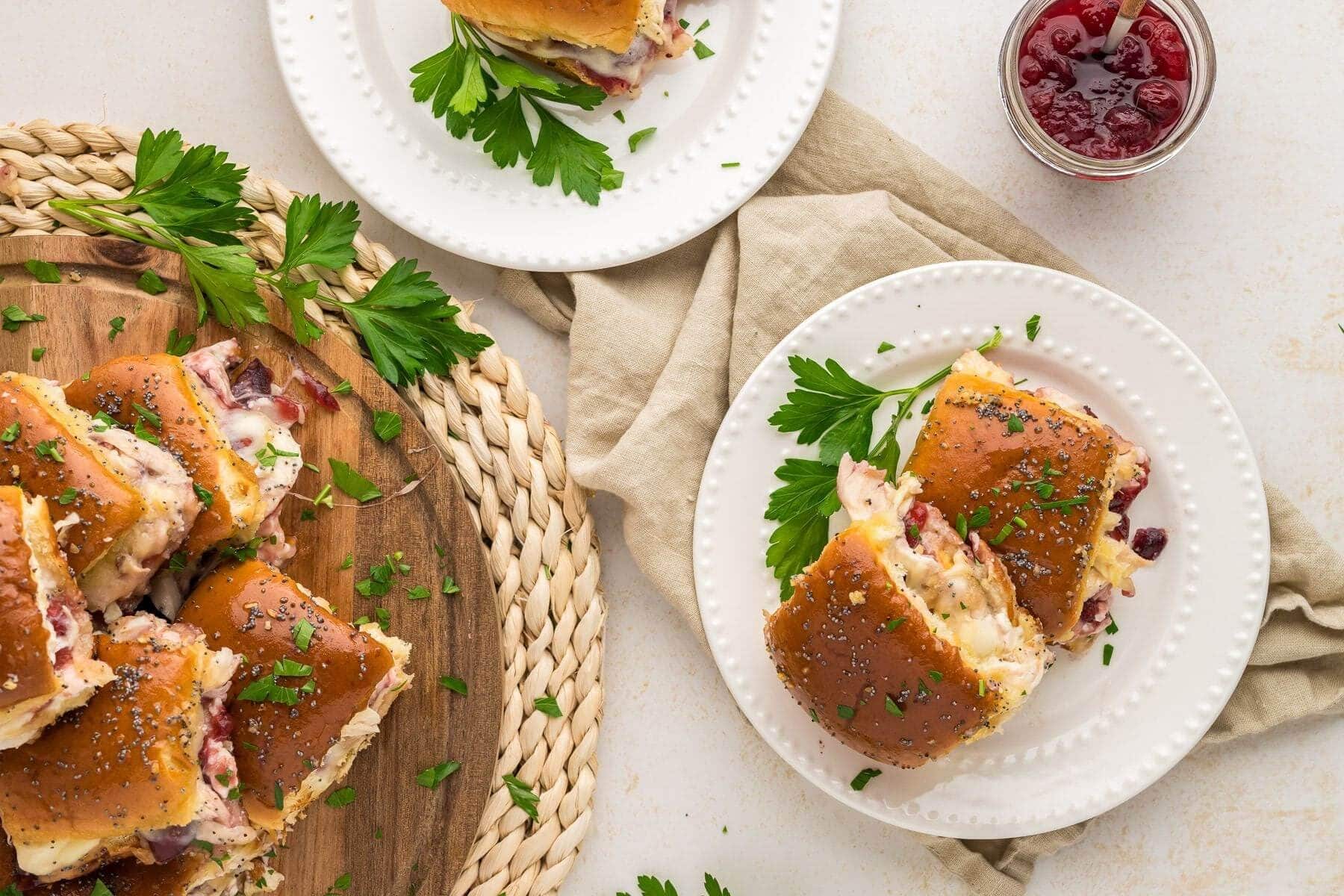 Two plates shown with easy baked turkey sliders on Hawaiian rolls next to the wooden board with piled up sliders.
