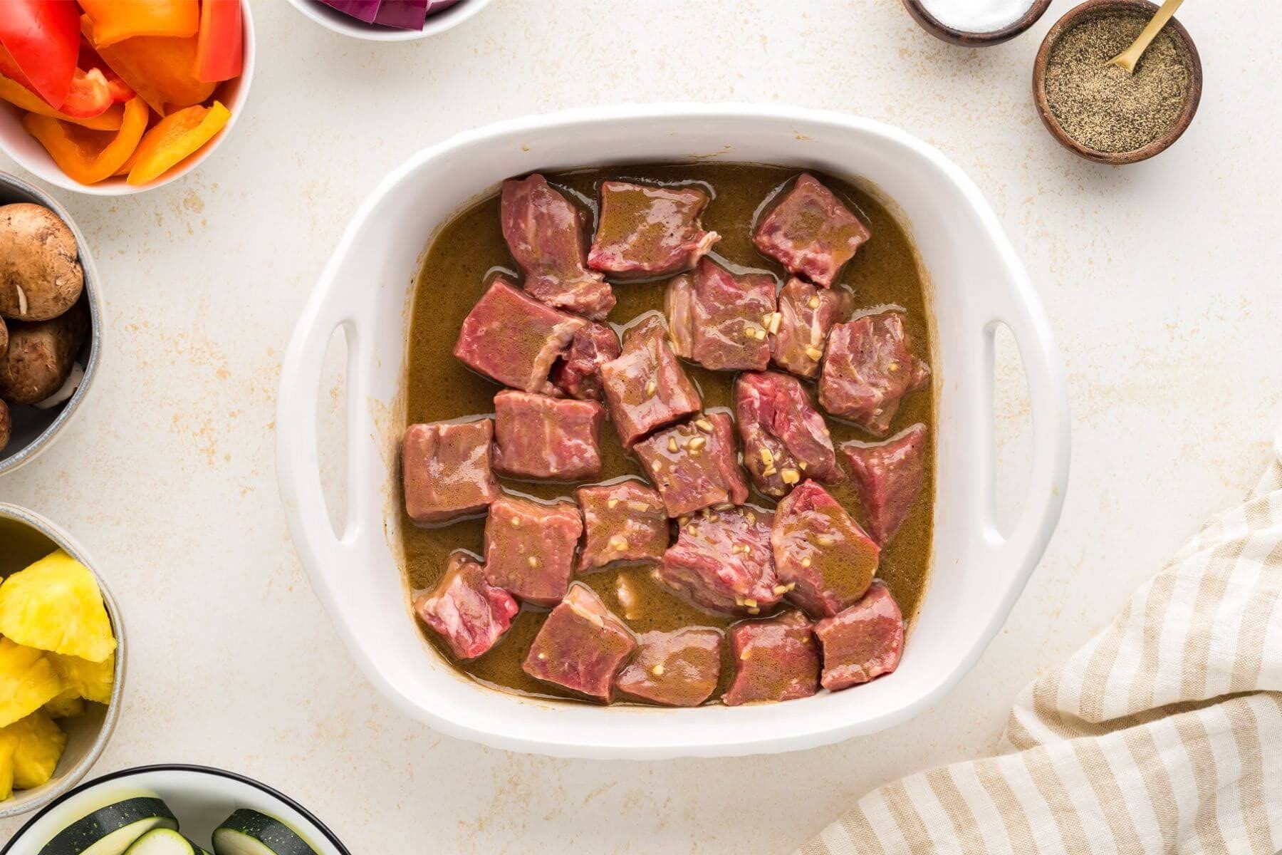 Casserole dish with steak marinating in shish kabab marinade with veggies in background.