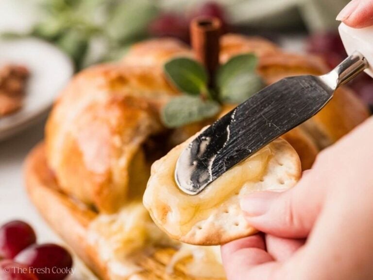 Spreading melted brie onto a cracker with baked pumpkin shaped brie in background.