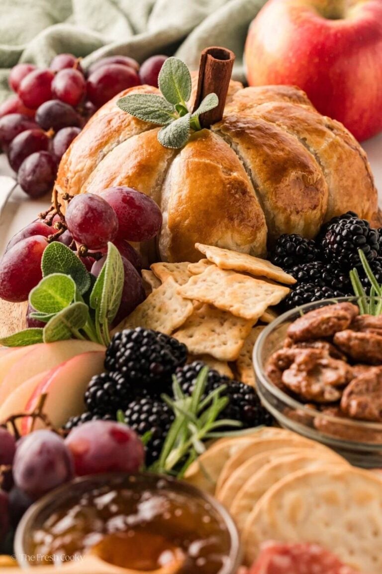 Golden, brown and flaky baked pumpkin shaped brie on board with red grapes, berries, crackers, sage, nuts, apples.