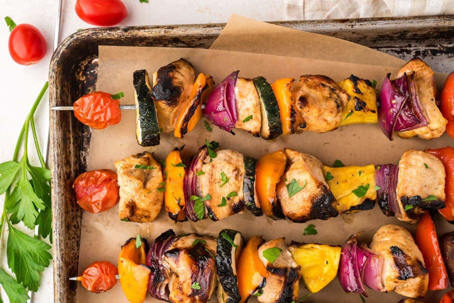 Three shish kabobs assembled vertically on a sheet pan bordered by small tomatoes.
