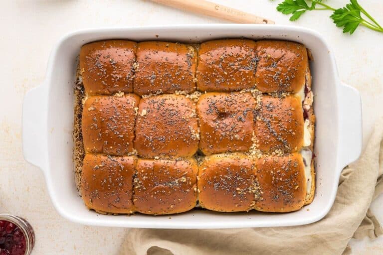 Turkey sliders in baking dish, topped with buns and brushed with butter sauce sprinkled with poppy seeds.